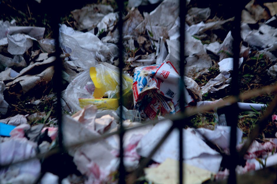 Close-up view of a pile of scattered waste materials and rubbish behind black metal railings, including crumpled paper, plastic bags, cardboard packaging, and food wrappers, on the ground outside a building. The waste appears to be discarded outdoors, with some debris partially obscured by the railings. This scene illustrates improper waste disposal in the context of environmental cleanliness and local waste management regulations, relevant to understanding waste rules in Lee. The photograph captures the mess and clutter typical of unmanaged rubbish accumulation, supporting informational content about lawful disposal practices involved in house removals and waste regulations referenced in the page about avoiding council fines.