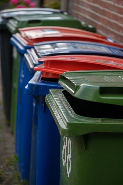A row of nine large wheeled wheelie bins lined up along a pavement adjacent to a brick wall in an outdoor setting. The bins are made of durable plastic and each has a hinged lid, with colors including green, blue, red, and black, indicating different waste categories. The green bin in the foreground is partially open, revealing an empty interior. Behind it, the blue and red bins are closed, and the remaining bins in the row are also closed, with their lids securely in place. The bins are positioned closely together, suggesting they are placed for regular waste disposal or recycling collection. The environment appears well maintained, with natural daylight illuminating the scene, capturing a typical outdoor waste collection area that would be encountered during house relocations or waste disposal in Lee. The presence of [COMPANY_NAME] and the reference to house removals or packing processes is relevant for understanding waste management rules during moving services.