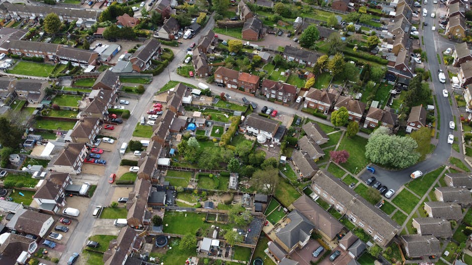 Aerial view of a residential neighbourhood showing a dense arrangement of houses with well-maintained gardens and backyards, some featuring patios, sheds, and garden furniture. Cars are parked along the narrow streets that wind through the estate, with a mix of family homes, terraced houses, and green spaces with trees and lawns. In the foreground, a removal team from Man with Van Lee is seen unloading boxes and furniture from a moving van parked on the pavement, with some items wrapped in protective packaging like cardboard, plastic wrap, and furniture blankets. The team is engaged in the loading process, carrying items such as cardboard boxes, small household appliances, and pieces of furniture towards or away from the van. The scene is set during daylight with natural lighting, clearly illustrating the home relocation and furniture transport activities involved in house removals within the Horn Park Estate area, emphasizing professional packing, loading, and transport procedures.