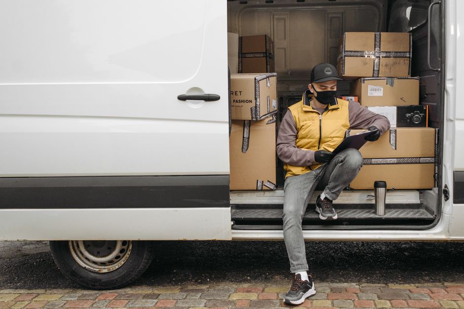 A man with a beard, wearing a black cap and a white T-shirt, seated inside a white moving van with the door open, is holding a smartphone with both hands. The interior of the van, visible behind him,