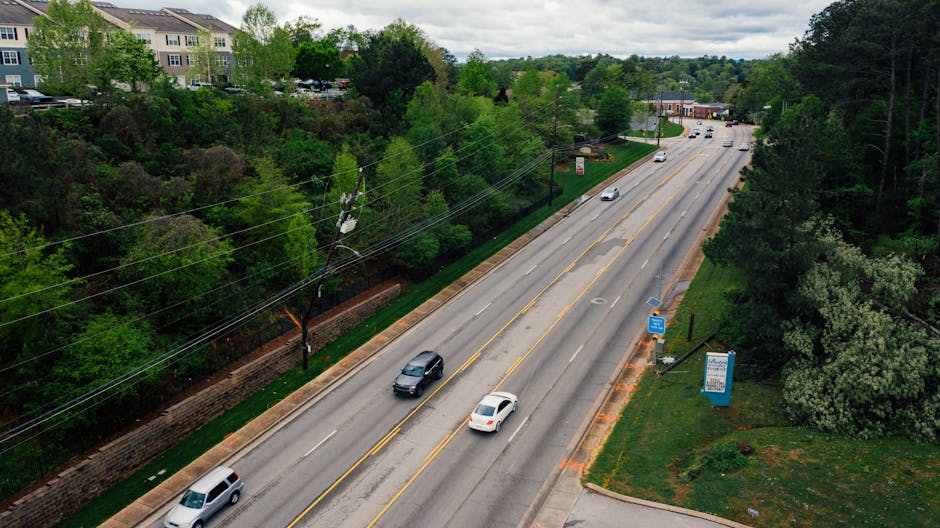 An aerial view of a multi-lane road flanked by greenery and trees, with a low brick wall and residential buildings visible on the left side. Several cars are seen travelling in both directions, with a few waiting at a traffic light. The road features painted lane markings, including a turn lane, and has a few vehicles parked near the pavement on the right. The weather appears cloudy, and the area is well-maintained with grass and bushes alongside the pavement. This scene relates to house removals and moving logistics, illustrating a typical urban street environment suitable for loading and transport of furniture or packing materials often involved in home relocation services offered by companies like Man with Van Lee.