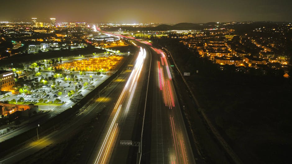 Aerial night view of a busy motorway near London, shown with long exposure light trails from moving vehicles in white and red across multiple lanes. To the left, there is a brightly lit parking lot with rows of cars parked among green trees and illuminated buildings. The surrounding area includes residential and commercial buildings with many lights turned on, visible on the right and in the background, extending towards a distant cityscape and hills under a dark sky. The image captures the urban environment at night, illustrating the infrastructure and traffic involved in home relocations and moving logistics, as seen by Man with Van Lee on their service page about best times to move and parking tips, highlighting the importance of effective planning for furniture transport and loading process during house removals.