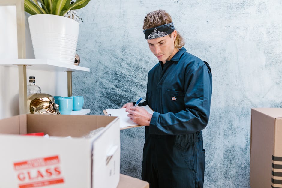 A man wearing a black cap, white polo shirt, and blue jeans is kneeling on the pavement outside a property, surrounded by cardboard boxes of various sizes, some open and some sealed with packing tape.