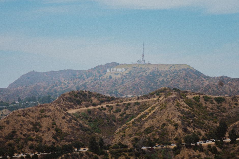 A daytime view of the Hollywood Hills hillside in Los Angeles, featuring dry, brown, and green vegetation covering the rolling terrain. Prominently visible in the distance is the iconic Hollywood sign, with large white letters spelling 'HOLLYWOOD' situated on the hillside. The sky above is clear with a few wispy clouds, and there is a tall radio or television antenna on a peak near the Hollywood sign. In the foreground, the lower hills are also covered with similar vegetation, and a line of trees can be seen near the base. This scenic landscape is often associated with the entertainment industry and celebrity culture. When considering house removals or home relocation services with Man with Van Lee, understanding the landscape and terrain of Los Angeles can assist in planning furniture transport and moving logistics through hilly or urban areas.
