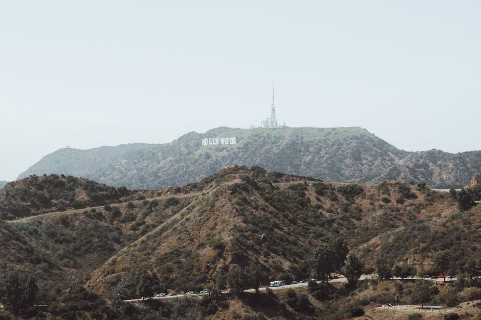 A wide view of the Hollywood Hills in Los Angeles, featuring the iconic Hollywood Sign prominently situated on a hillside covered with sparse vegetation and rugged terrain. The image captures multiple rolling hills with dry grass, small shrubs, and scattered trees. In the foreground, a winding road with vehicles passing along it is visible, running through the lower part of the image. The sky above is overcast, creating diffuse lighting over the landscape. This scene exemplifies the typical natural environment associated with Los Angeles, with the Hollywood Sign serving as a well-known landmark in the area. The image contextually relates to home relocation or moving services in the Los Angeles region, where viewing the landscape along with urban infrastructure supports understanding of local moving logistics, as found on websites like manwithvanlee.co.uk, which offers removals and furniture transport services in the area.