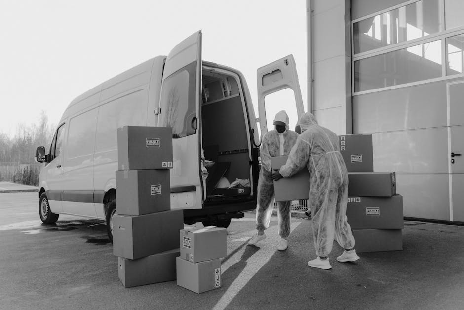 Two movers dressed in protective coveralls, gloves, and masks are unloading cardboard boxes from the open rear of a white van parked outside a modern building with large glass windows. The boxes vary in size and are being carried and stacked on the pavement as part of a home relocation process. The vehicle is positioned next to the building's entrance, suggesting they are in the loading or unloading stage of a furniture transport operation. The background includes an outdoor setting with clear skies and distant trees, indicating daylight conditions. The scene captures the practical steps involved in packing and moving, with the movers carefully handling the boxes during a quick, same-day removal service as provided by Man with Van Lee, which specialises in urgent removals and efficient logistics for house removals.