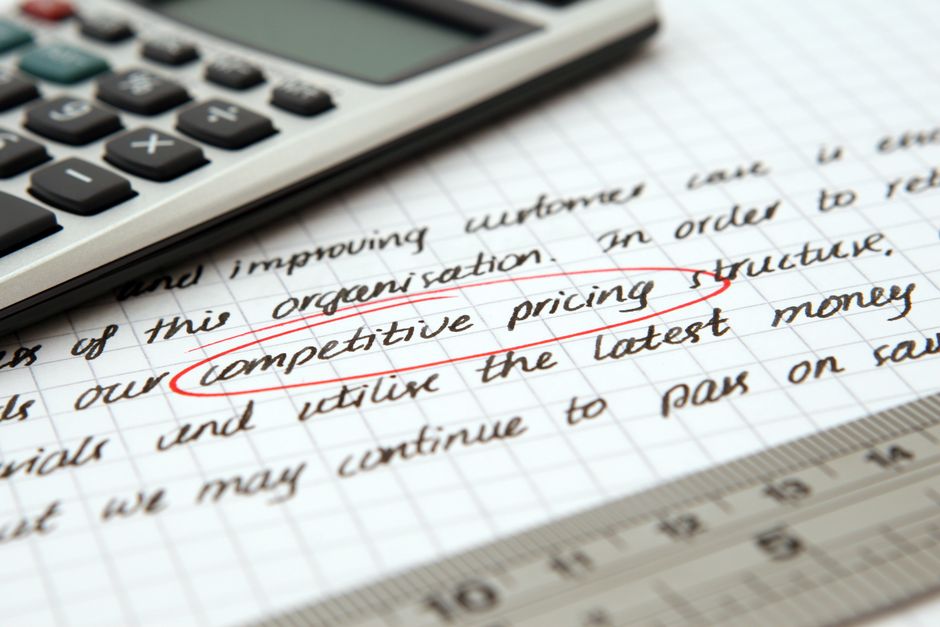 Close-up image of a sheet of graph paper with handwritten notes about competitive pricing structures, featuring the phrase 'competitive pricing structure' underlined in red. A visible calculator with black and white buttons is partially placed on the paper, suggesting calculations related to moving costs. The background includes a metallic ruler along the bottom edge and a glimpse of a transparent plastic folder. The scene indicates a focus on financial planning or cost analysis, relevant to house removals or moving services, as part of the context of transparent quotes and moving logistics discussed on the page from Man with Van Lee.